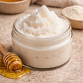 Open glass jar of Cashmere sugar scrub with a soft white, fine sugar texture, placed on a folded cashmere sweater with a wooden honey dipper nearby.