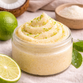 Open glass jar of Coconut and Lime sugar scrub with a soft yellow, fine sugar texture, topped with lime zest and placed on a neutral surface with fresh lime and coconut nearby.