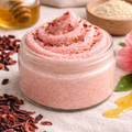 Open glass jar of Hibiscus and Honey sugar scrub with a soft pink, whipped texture, topped with dried hibiscus petals, placed on a light fabric surface with honey and hibiscus flowers nearby.