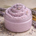 Open glass jar of Lavender & Vanilla sugar scrub with a light purple whipped texture, surrounded by dried lavender on a stone surface with a gold lid nearby.