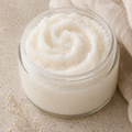 A clear glass jar filled with white exfoliating scrub, set on a textured beige surface with a draped beige linen cloth and small dried flowers.